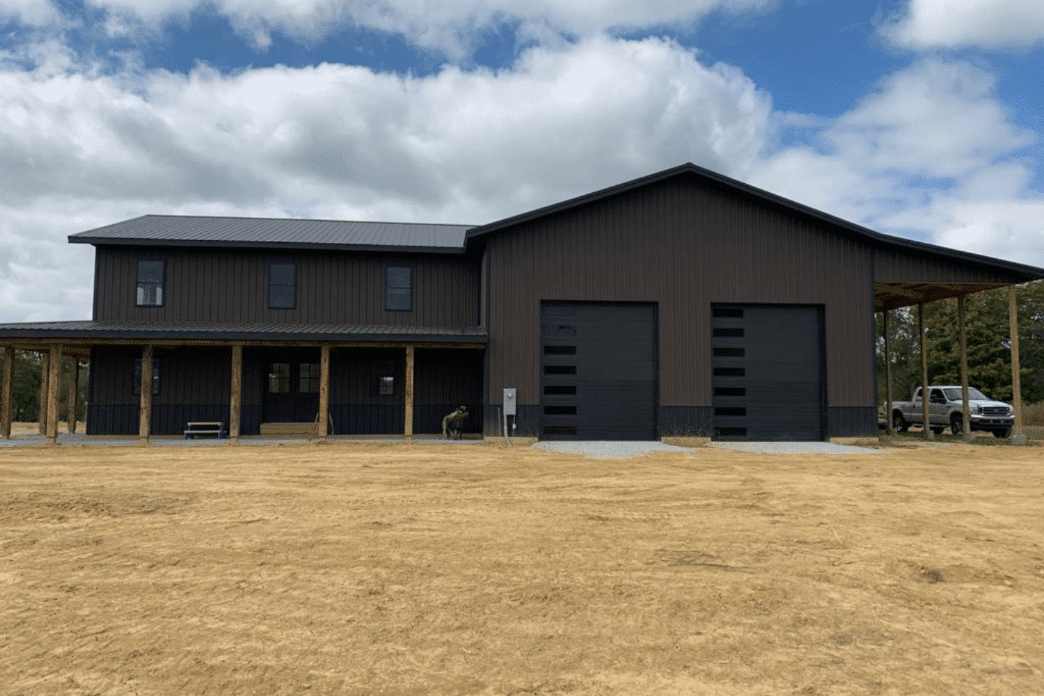 A large 30x100 barndominium kit with a dark brown exterior and black trim, featuring a two-story living area on the left with a covered porch supported by wooden beams. The right side includes two large black garage doors and an extended open-sided structure. The building is surrounded by a dirt lot under a partly cloudy sky with a truck parked to the side.