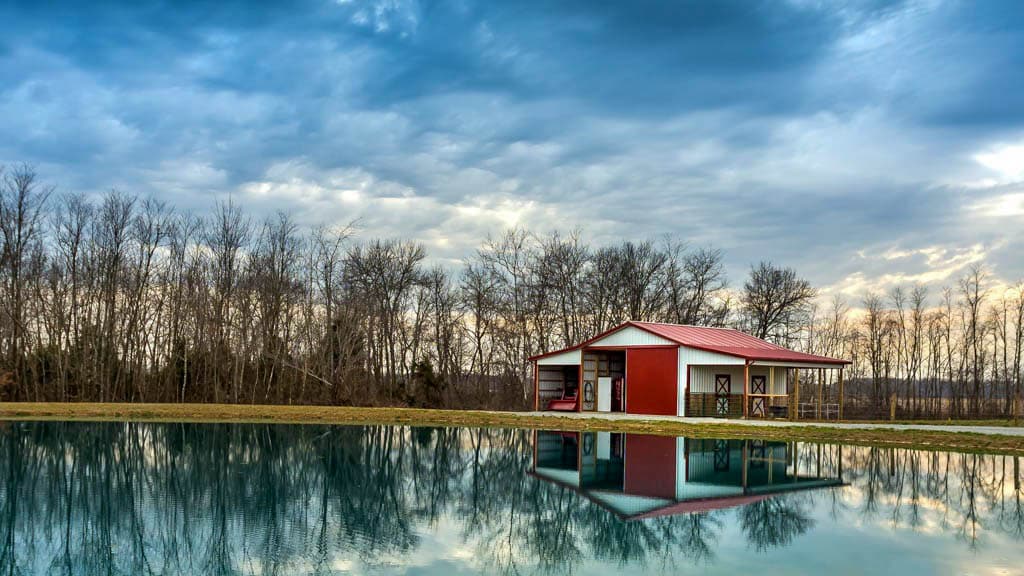 A 30x40 equestrian pole barn with a red roof standing right by a lake