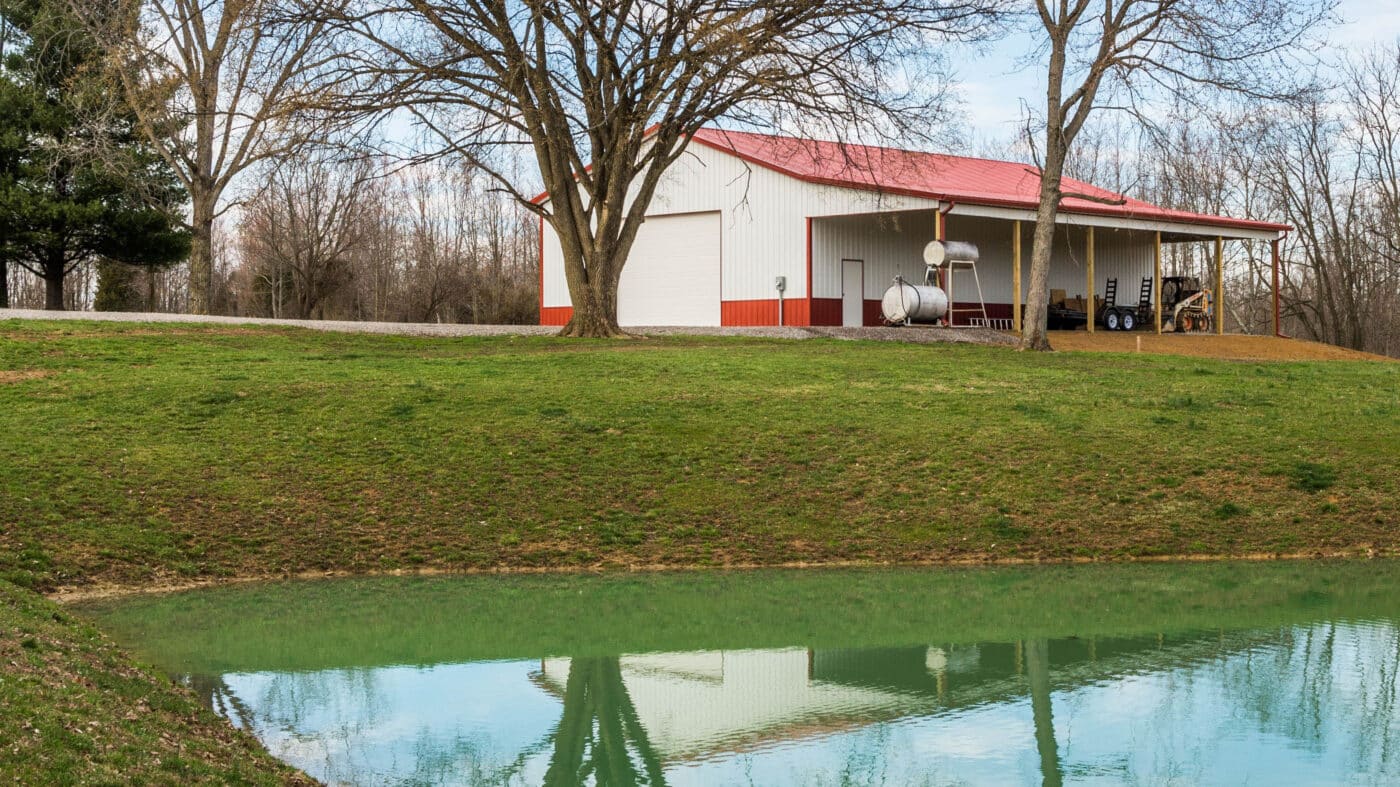40x64-agricultural-pole-barn-with-white-siding-red-trim-a-red-metal-roof-and-a-white-garage-door-with-farming-equipment-under-a-front-porch-by-a body of water