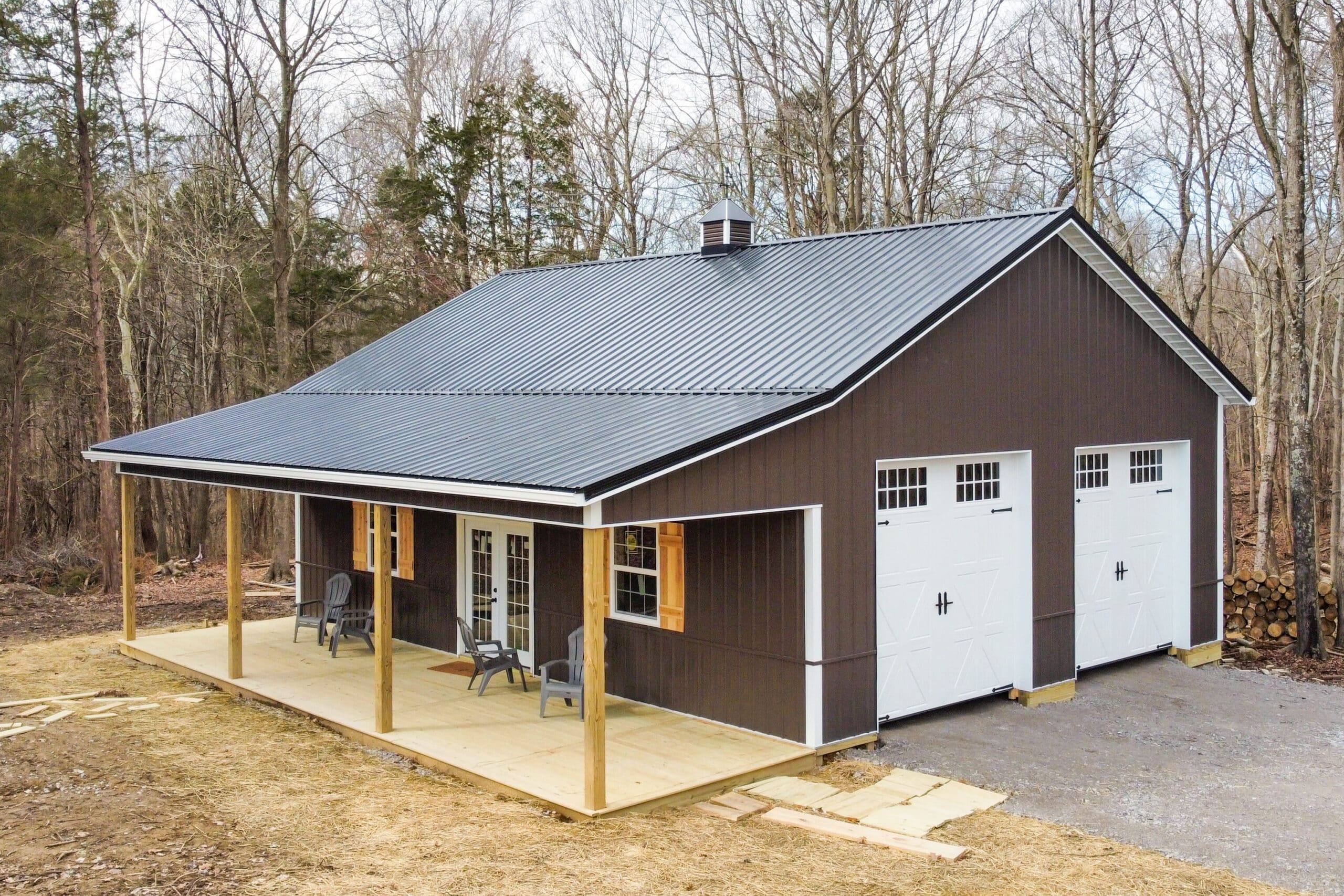 brown pole barn garage with black metal roof sweeping down into overhang over concrete patio featuring two white carriage garage doors on the left side