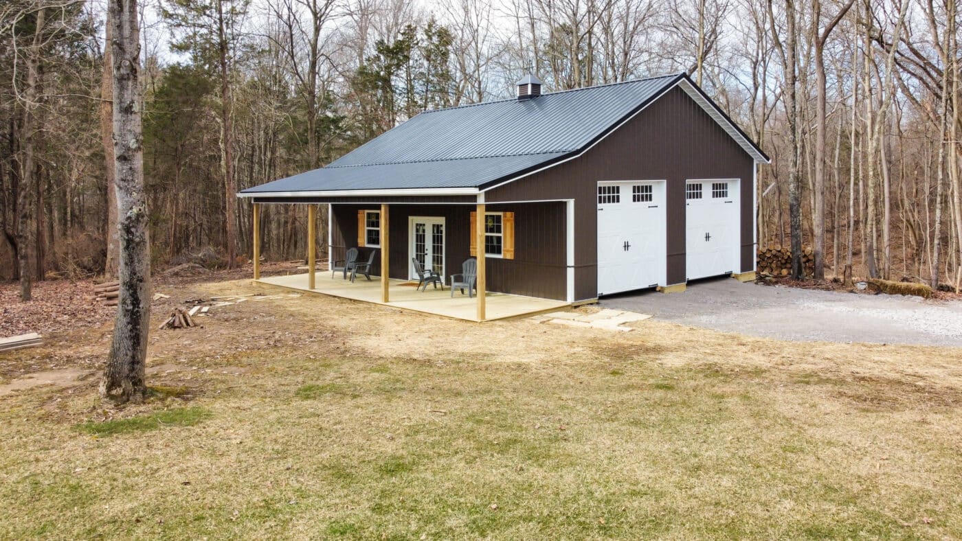 brown pole barn garage with two white overhead doors on the side with transom windows and a black metal roof featuring an overhang over a concrete patio with chairs on it