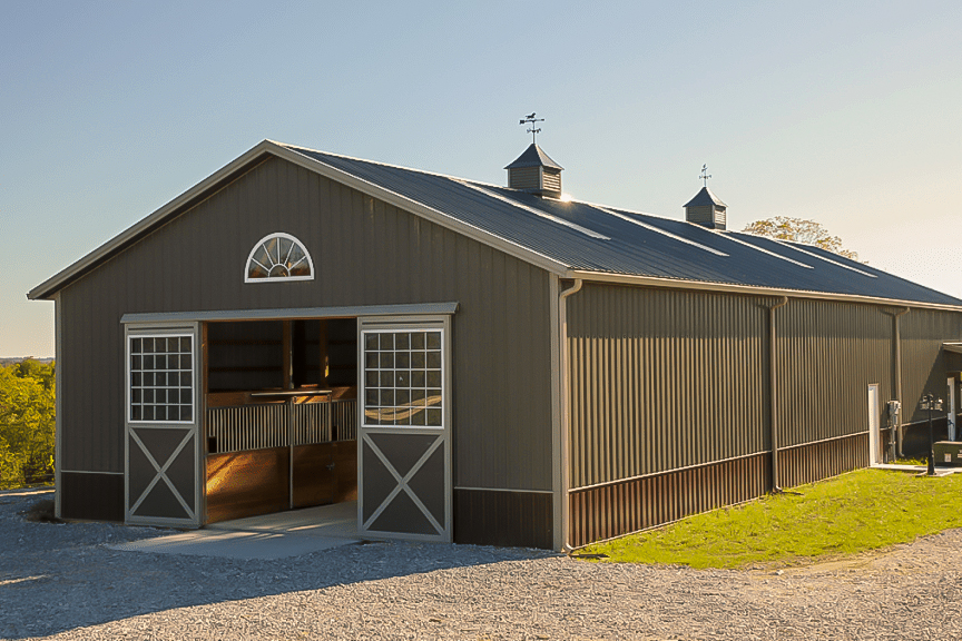 gray agricultural pole barn with brown metal paneling along bottom and black metal roof with two cupolas featuring open slider doors showing multiple wooden stalls inside