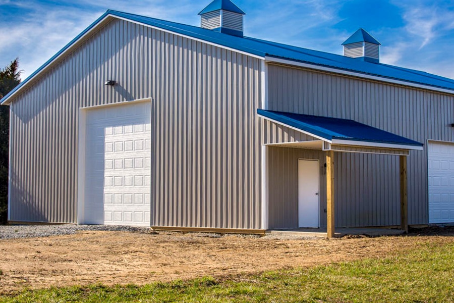 gray and dark blue roofed pole barn with two cupolas two overhead doors and side door with overhang