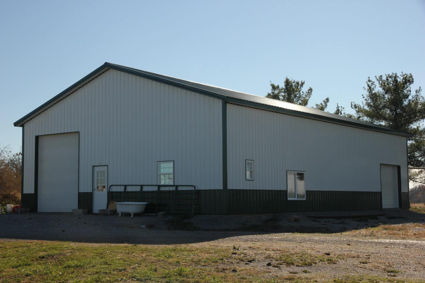 light gray pole barn garage with dark gray trim and metal roofing and two white overhead doors on on the front and one on the side featuring windows of multiple sizes and styles