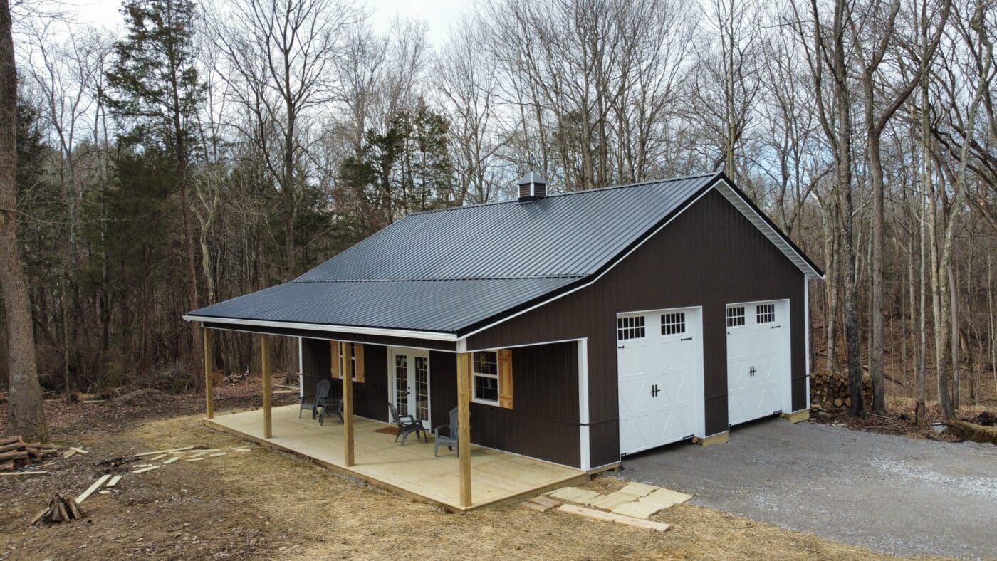 brown pole barn garage with white trim, a porch, black roof, and two white garage doors in McArthur OH