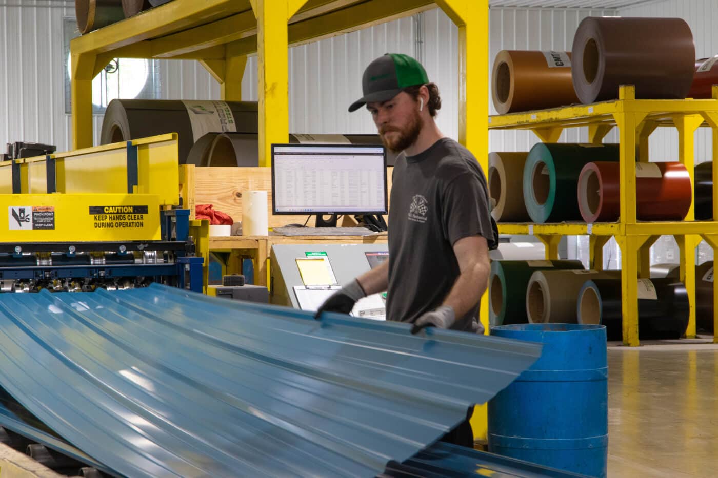 A worker in a green cap and black shirt operates machinery to produce blue metal panels inside a manufacturing facility. The large yellow machine processes the panels, while rolls of raw metal and a computer station are visible in the background.