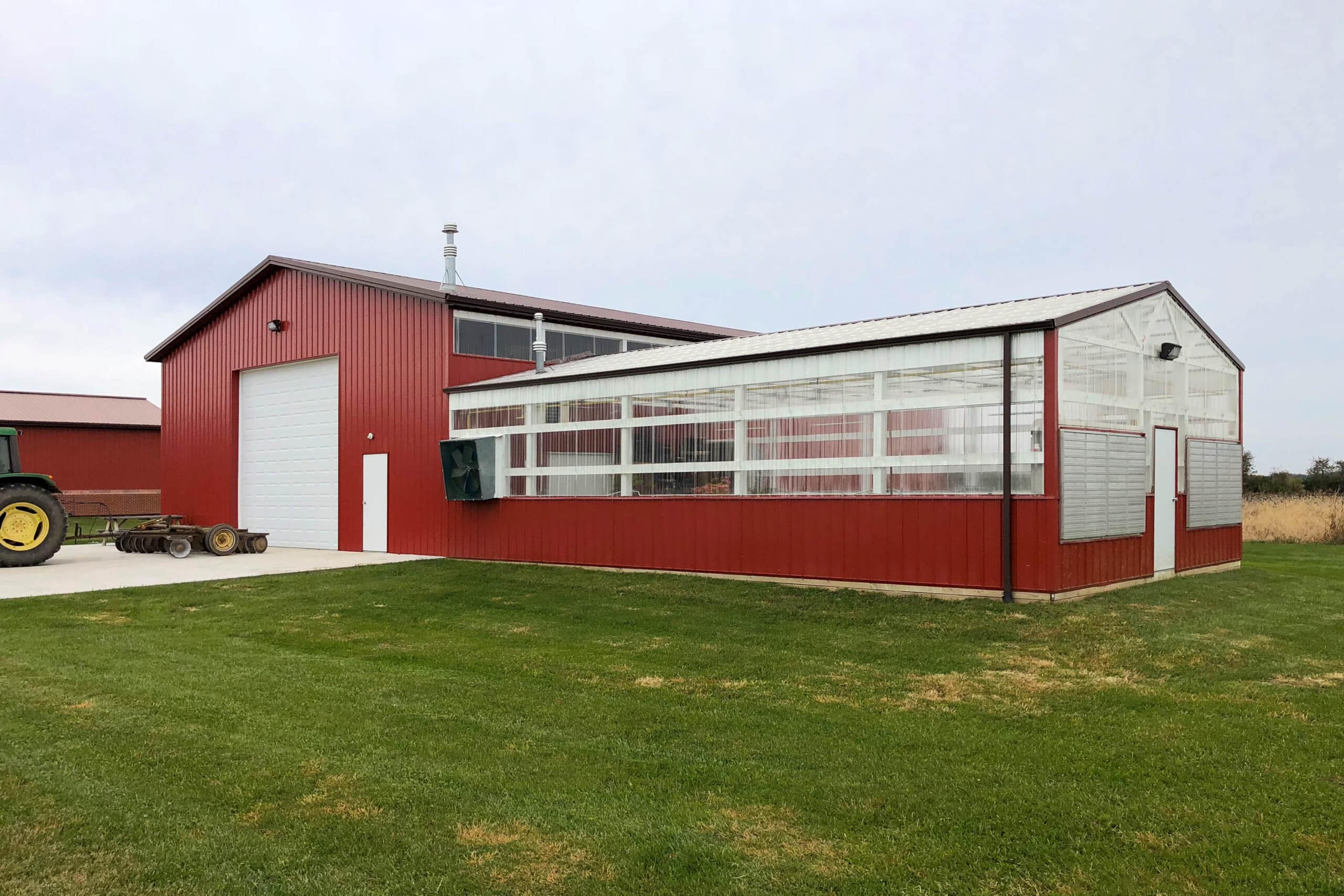 red metal sided 30x70 agricultural pole barn with clera green house siding as well featuring one white overhead door with tractor parked in front
