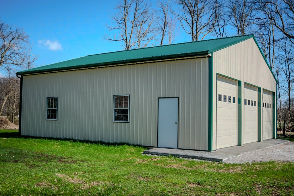 side view of 30x50 pole barn garage with cream colored metal siding green trim and metal roofing as well as three white garage doors with transom windows a white side door and two windows