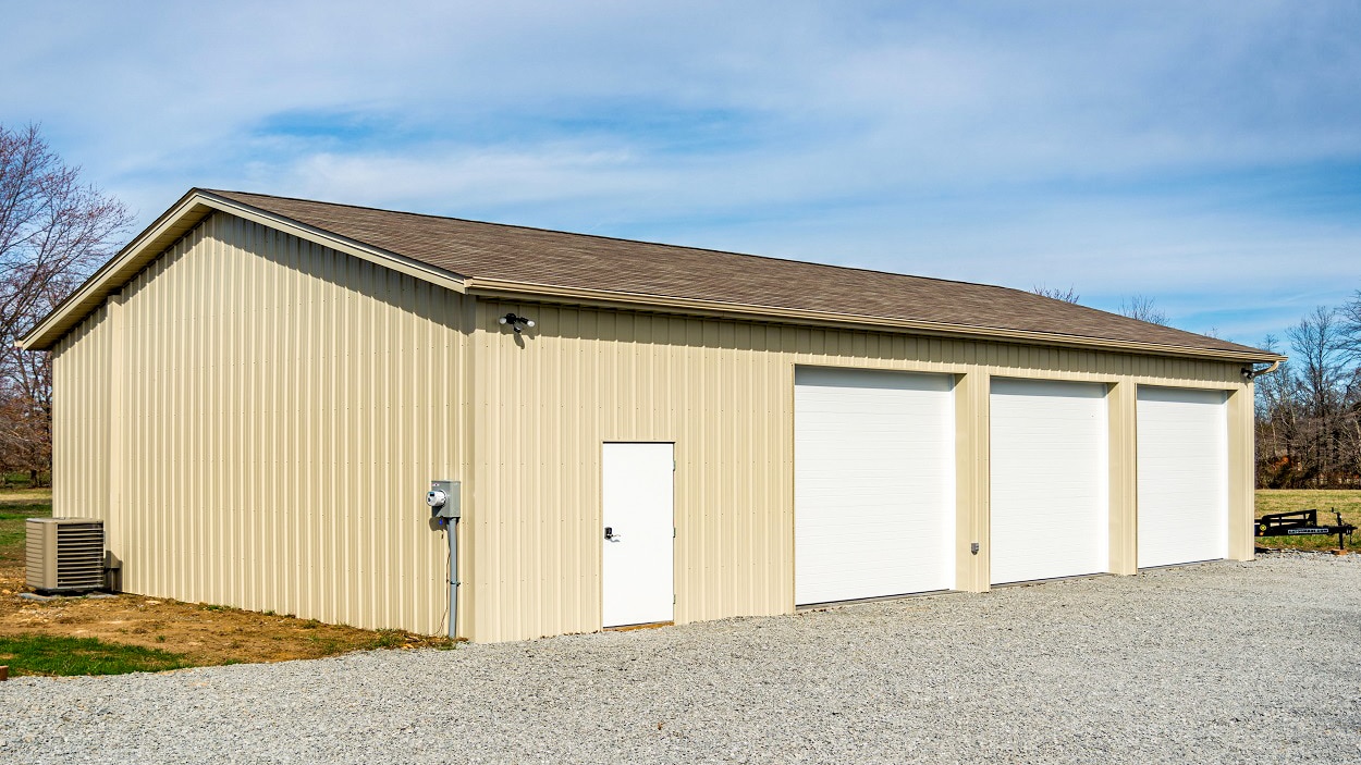 light yellow 80x90 pole barn garage with brown shingle roof three white overhead doors and a white side door along gravel drive