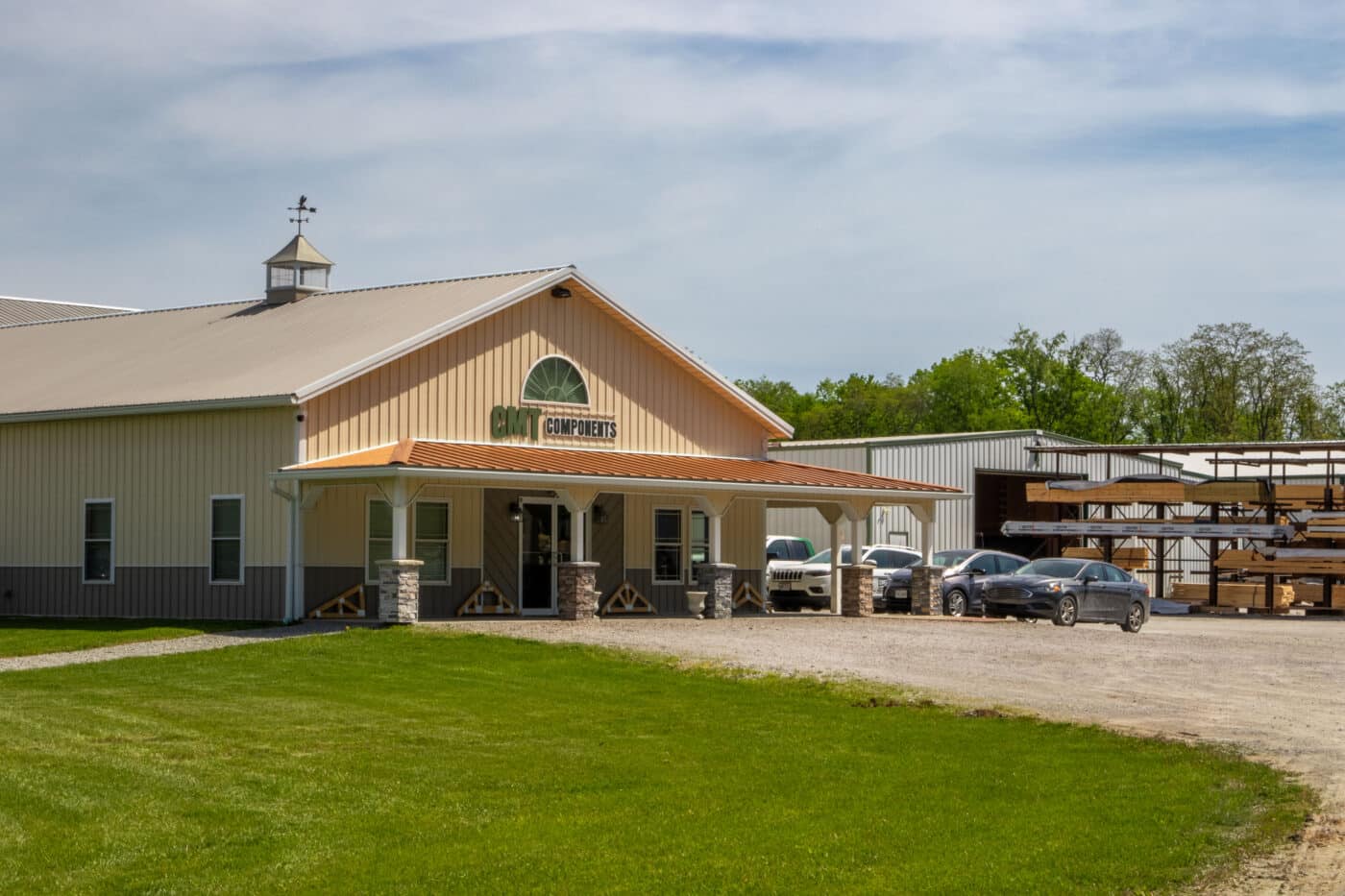 tan CMT components building with orange metal overhang and gray metal detailing along bottom with multiple cars parked in lot beside it