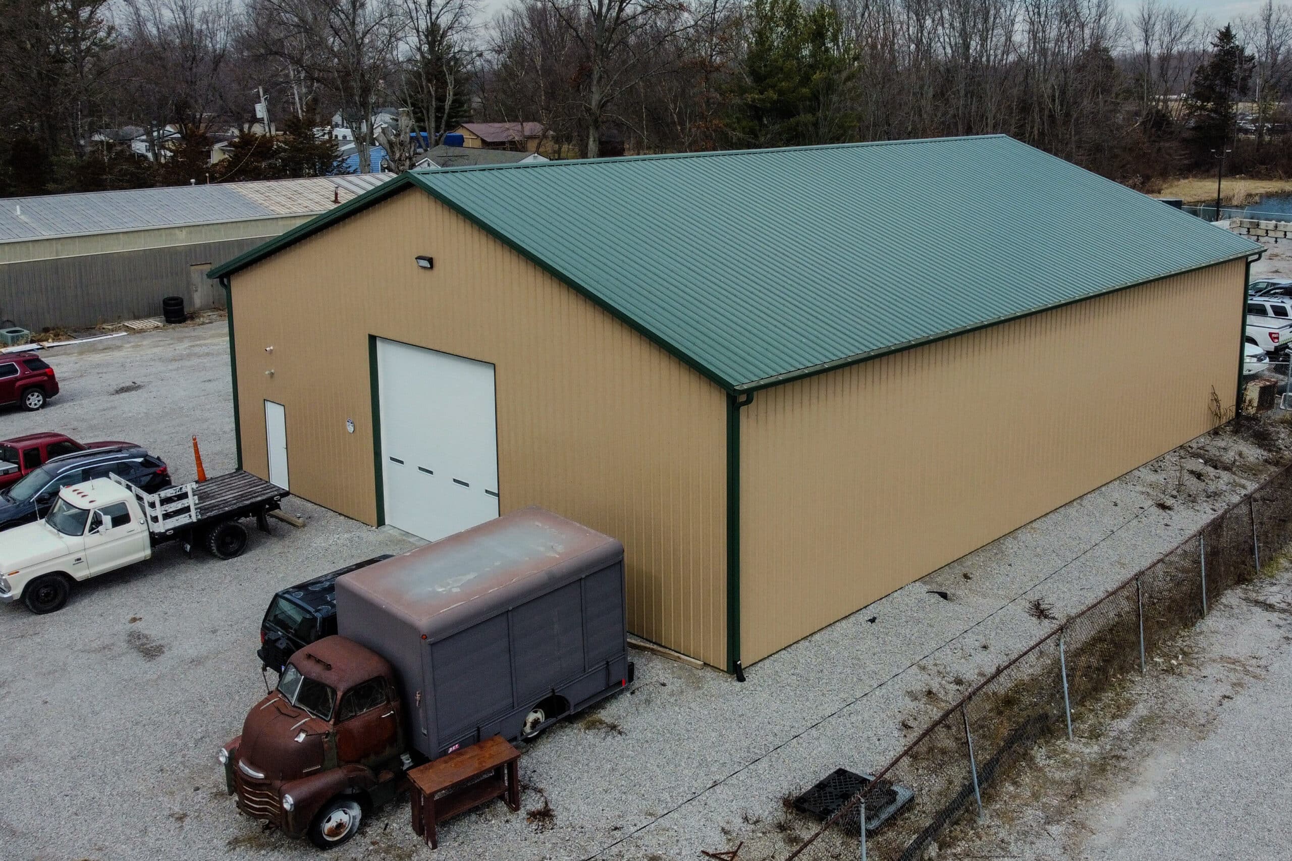 tan and green trimmed metal pole barn garage with green metal roof featuring white overhead door and multiple vehicles in yard around it