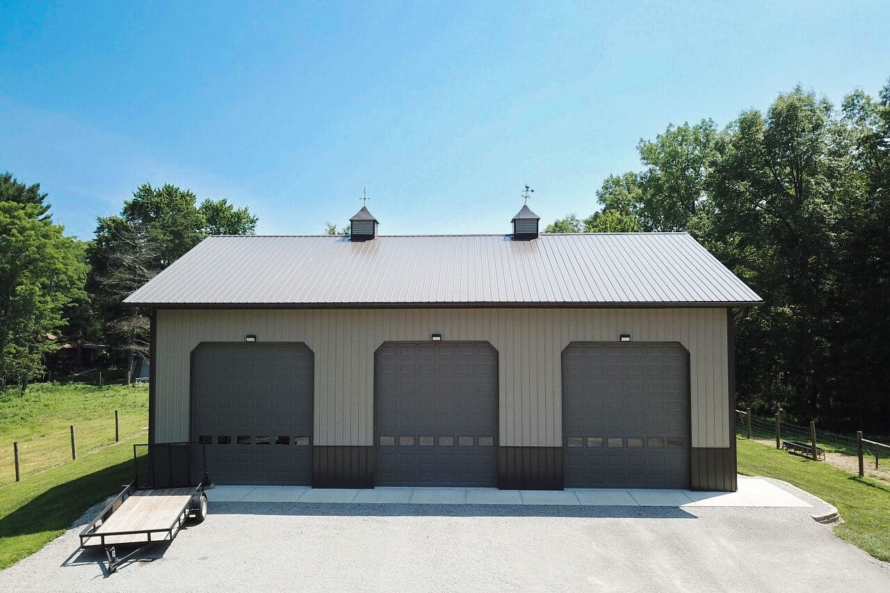 tan pole barn garage with dark brown metal roof and paneling along the bottom and three brown overhead garage doors
