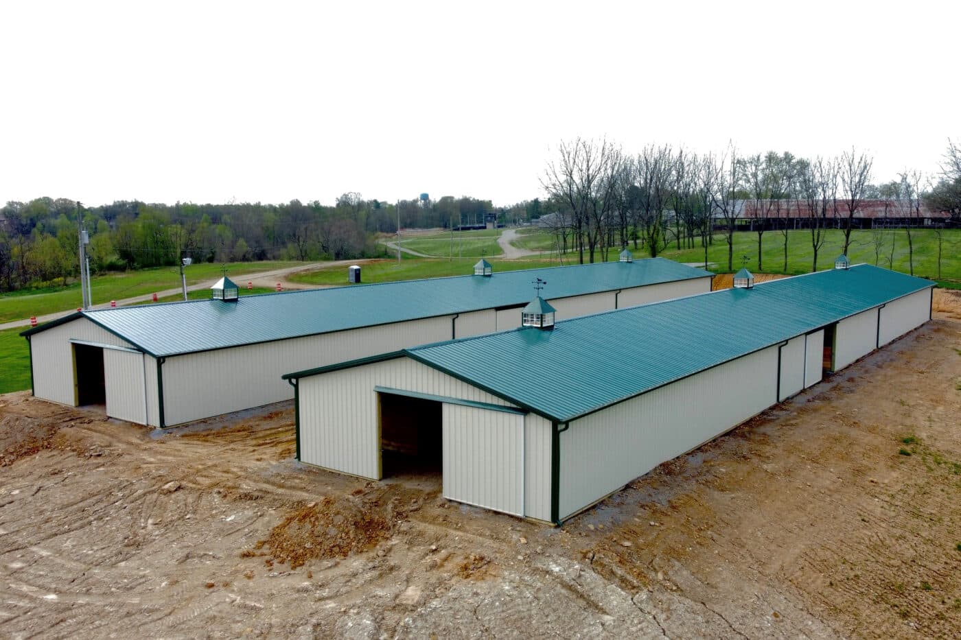 two tan agricultural pole barns with slider doors green trim and green metal roofs and three cupolas