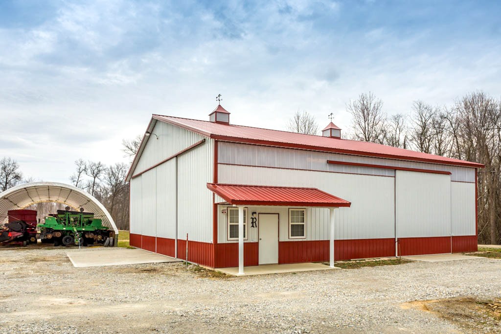 white and red trimmed agricultural pole barn with side overhang over white side door slider doors and outdoor shelter for equipment next to it