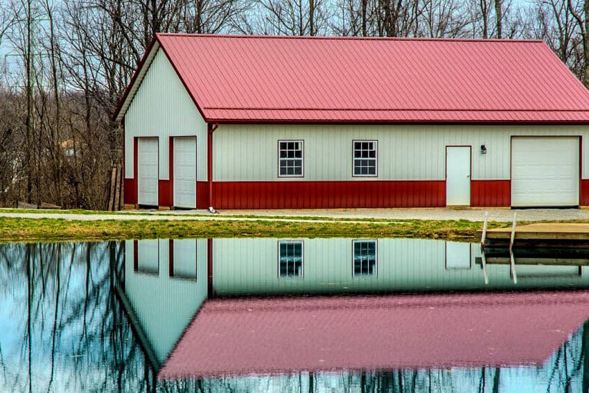 white pole barn with red metal trim on bottom and corners and a red metal roof featuring one white overhead door and regular door and two windows on the front and two white side overhead doors with the entire structure overlooking a body of water