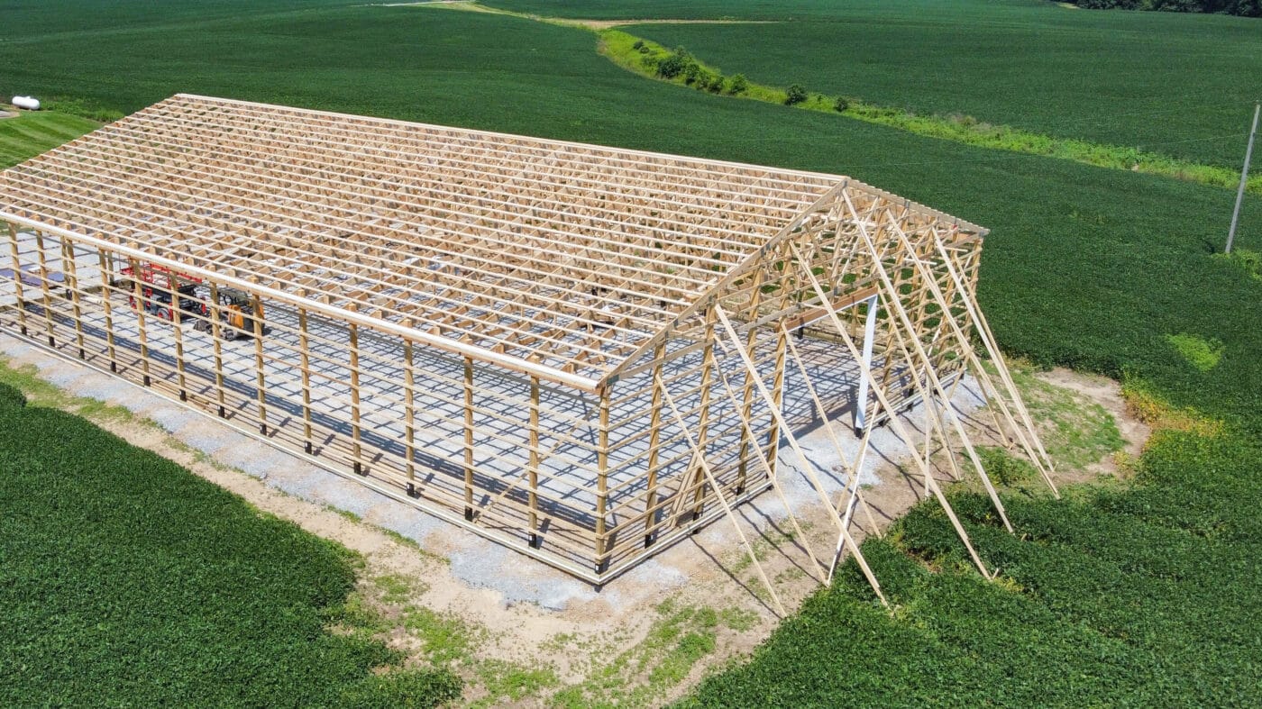 wooden pole barn frame in back of brick house on long gravel drive surrounded by green fields