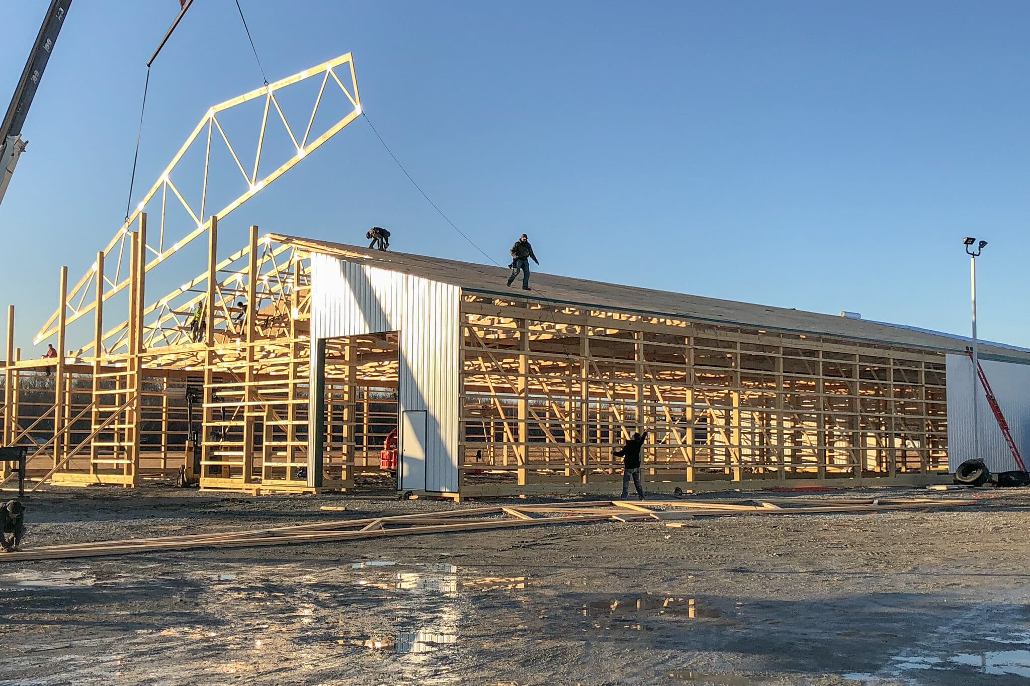 wooden roof truss being lowered onto frame of pole barn with white metal siding partially on it by crane with group of men on roof to help install it